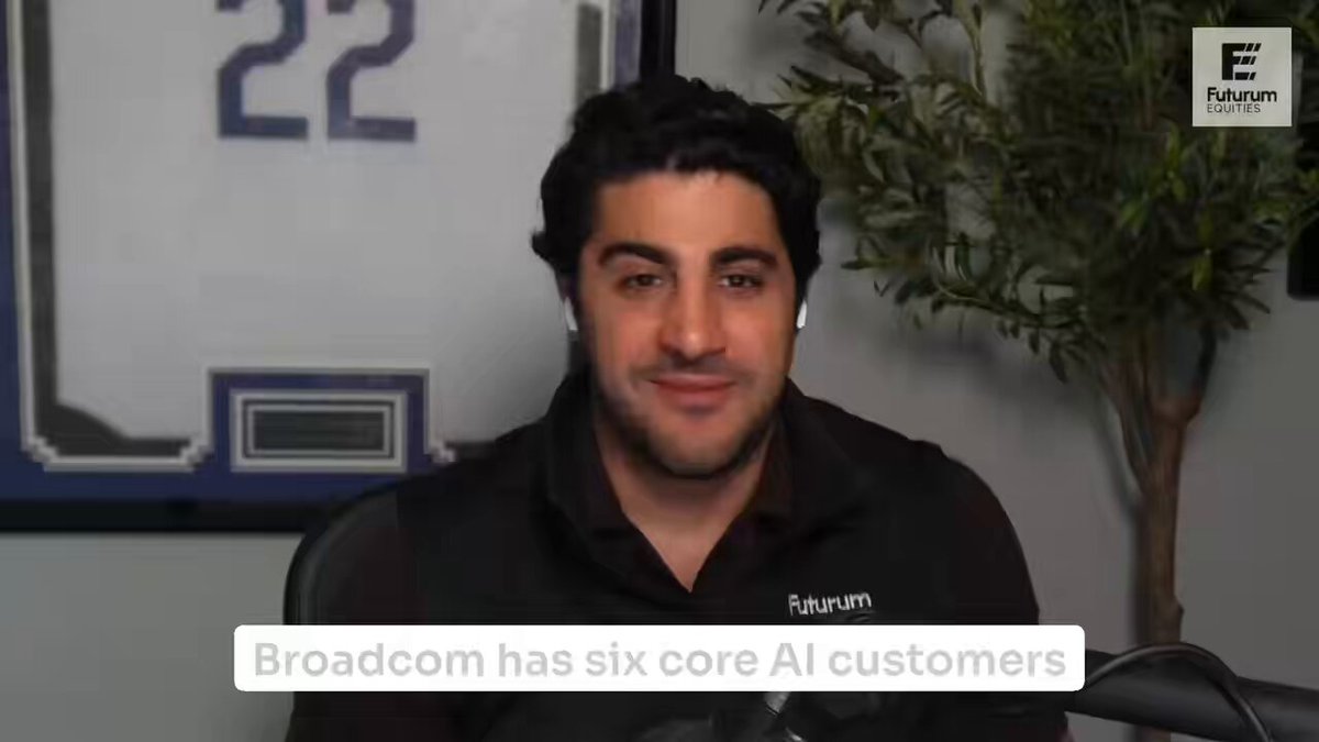 The image shows an interview with a man wearing a black shirt, sitting at a desk or chair. The man appears to be smiling as he speaks into a microphone. In front of him, there are two posters - one displaying a baseball player's name "AVGO" and another showing the word "FEW". These posters seem to be related to the interview topic.
