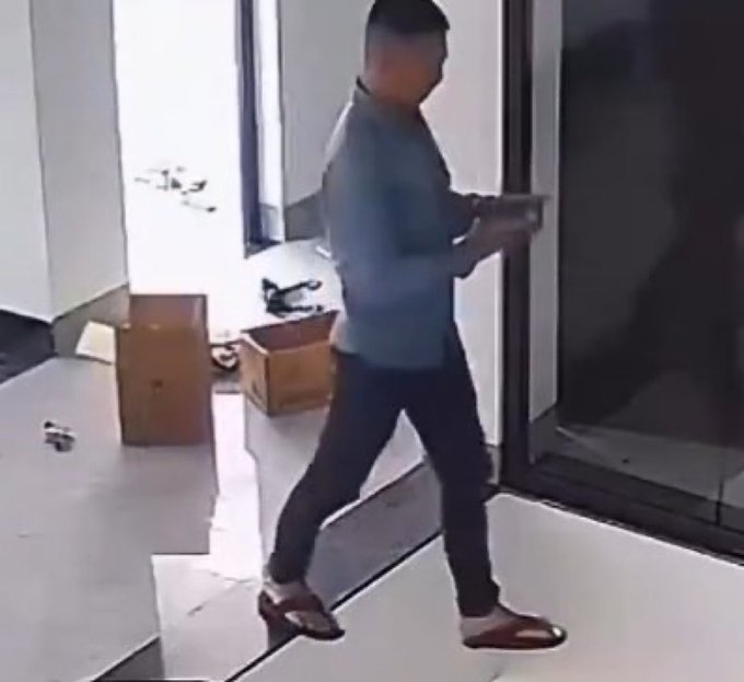 A low-resolution security camera capture showing a man in a grey shirt and dark pants walking through an indoor space carrying a box, with cardboard boxes and scattered debris on the floor.