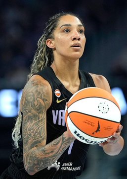 A female basketball player with braided hair and a tattooed arm prepares to shoot a WNBA basketball while wearing a black Valkyries jersey with Kaiser Permanente sponsorship.