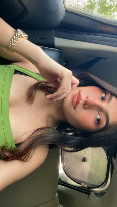 A woman taking a selfie in a car, wearing a green top and a gold watch, with makeup and styled hair visible.