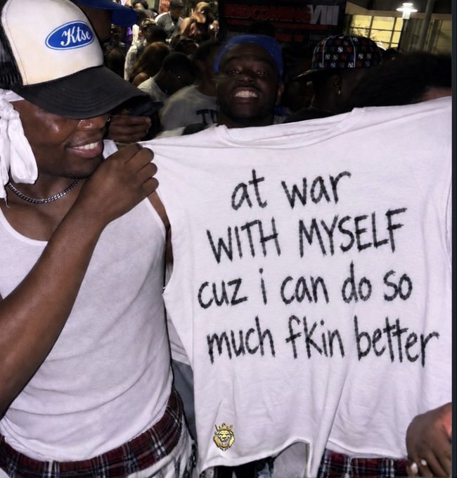 Two men smiling at a crowded concert; one holds up a white t-shirt with handwritten text reading 'at war WITH MYSELF cuz i can do so much fkin better' and a small yellow lion logo.
