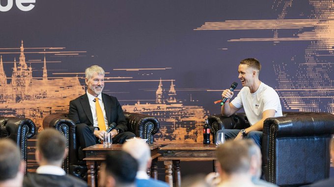 Two men seated on stage in leather armchairs at what appears to be a panel discussion, with one holding a microphone; the backdrop shows a stylized cityscape featuring a cathedral.