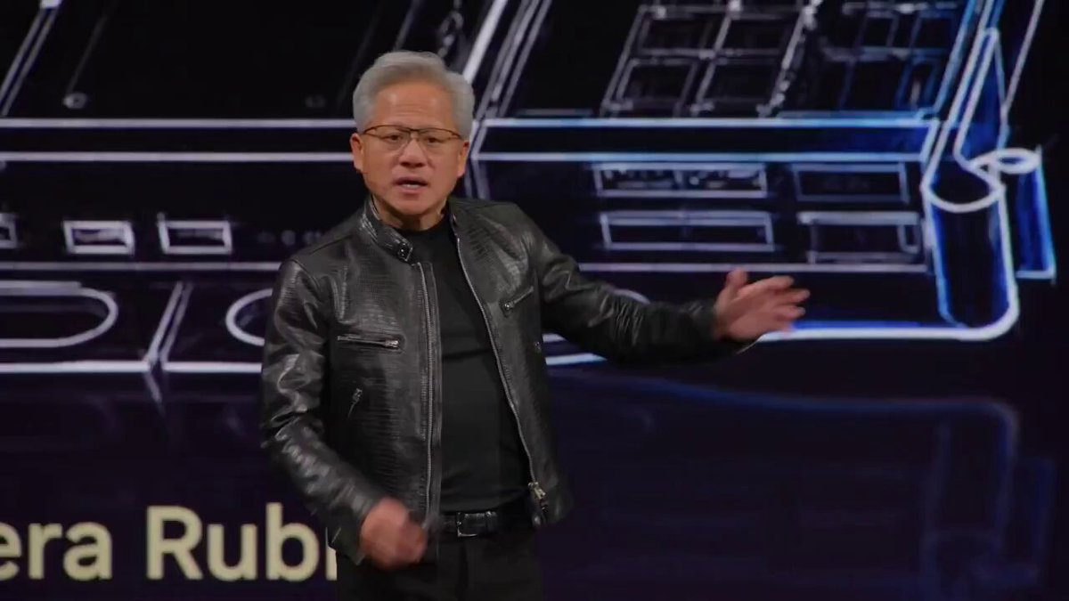 The image shows an older man wearing glasses standing in front of a computer screen with the words "LATEST: Nvidia $NVDA is building computers for AI agents." The man appears to be giving a speech or presentation, possibly at a conference or event related to the development and use of artificial intelligence.