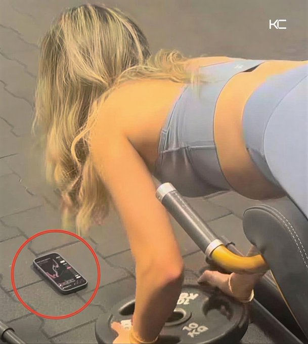 A woman exercising with a barbell, with her phone on the ground circled in red showing an active workout display.