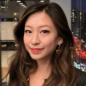 A close-up portrait of an East Asian woman with long brown hair, wearing red lipstick and dangling earrings, smiling against a nighttime city skyline backdrop.