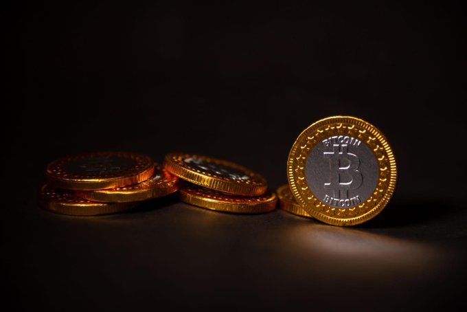 The image features a stack of gold coins with one coin prominently displaying the word "bitcoin" above its center. The other coins are positioned around this central coin, creating an interesting visual effect against a black background. The text in the image reads "Bitcoin starting to separate from the rest of the market again is becoming hard to ignore while BTC continues holding above its 200-week moving average". This implies that Bitcoin's value has been steadily increasing over time and is now outperforming other assets like ETH, SOL, and XRP.