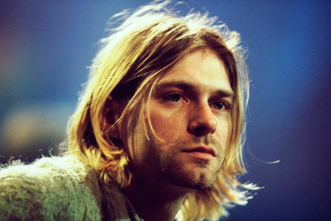 A close-up photograph of a man with long, tousled blonde hair and light stubble, wearing a textured cardigan, gazing intently with a soft, moody lighting against a dark blue background.