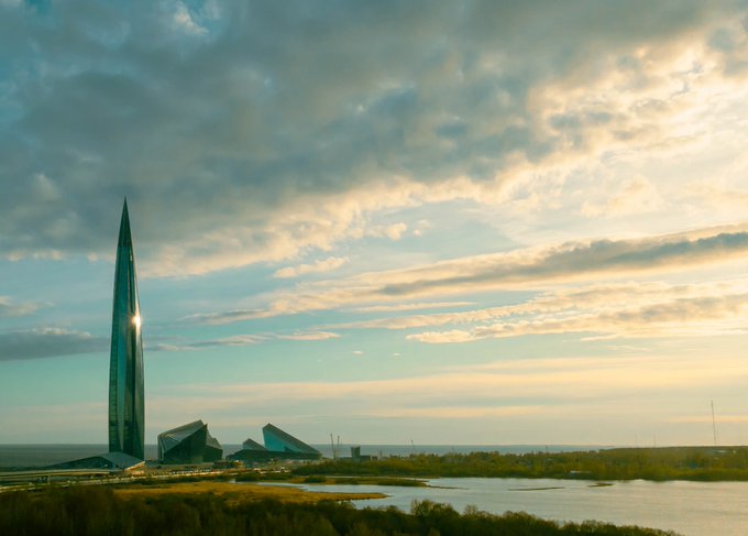 A sleek, tapered skyscraper and modern geometric structures rise from the shore of a calm body of water under a dramatic golden-hour sky, creating a striking cityscape silhouette.