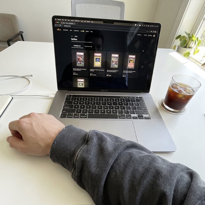 A person's arm in a gray hoodie rests on a desk next to an open MacBook Pro displaying a YouTube channel page, with a glass of iced coffee nearby and a plant in the background.