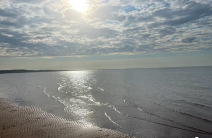 A serene beach scene featuring rippled sand in the foreground, gentle waves rolling in, and a bright sun shining through scattered clouds over the ocean.