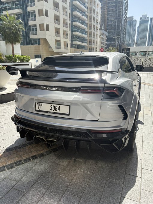 A silver Mansory-tuned Lamborghini Urus (Dubai plate U 3064) is parked in front of towering skyscrapers, showcasing the lavish lifestyle the "GM (get money)" caption celebrates.