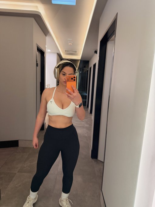 A mirror selfie in a hallway shows a woman wearing a white sports bra, black leggings, white sneakers, and headphones, holding an orange smartphone.