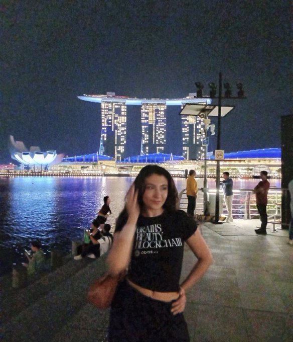 A woman poses at Marina Bay Sands in Singapore, with the iconic illuminated towers visible behind her across the water at night. Her shirt reads "Travis Scott Beauty Worldwide Astroworld," fitting the travel-focused post about her plans to attend Token 2049 Singapore and seek crowdfunding for her trip.