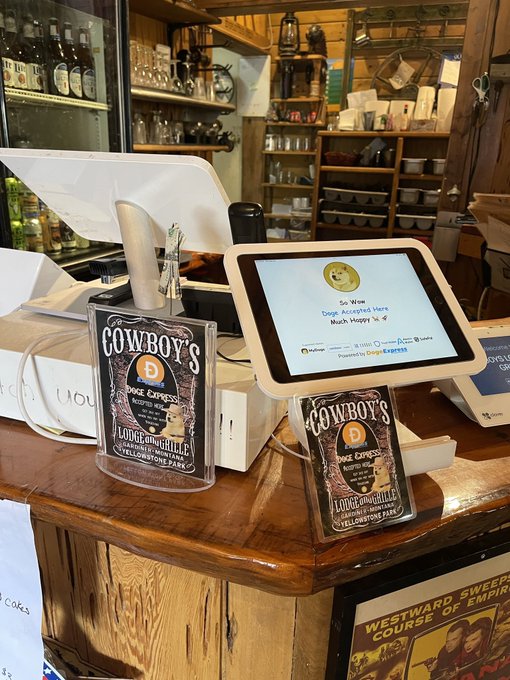 Two payment stands for "Cowboy's Lodge and Grille" sit on a wooden counter, each displaying a Dogecoin sign and a tablet screen prompting "So Now Dogs Accepted Here. Much Happy," illustrating the Gardiner, Montana restaurant's embrace of $DOGE payments.