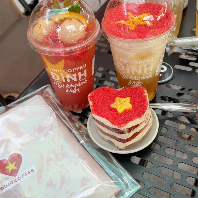 The image shows two drinks sitting on a table along with a cake decorated with red and white frosting that has a yellow star on top, possibly representing the Vietnamese flag. The background of the photo features a black metal grate or grill, which could be part of an outdoor dining area or a kitchen countertop.