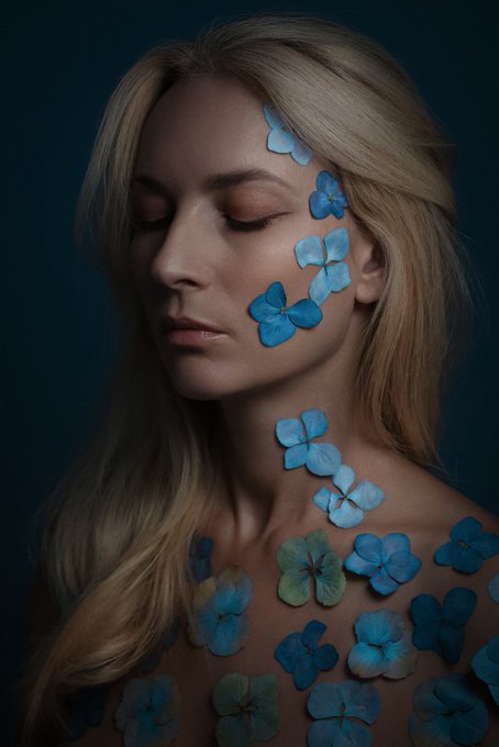 A blonde woman with closed eyes has delicate blue flowers arranged across her cheek, neck, and bare shoulder against a dark blue background.