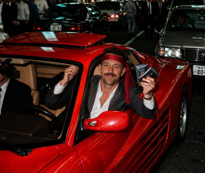 A man wearing a red headband sitting in the driver's seat of a red sports car, holding up trading cards, with Japanese license plates visible on nearby cars.
