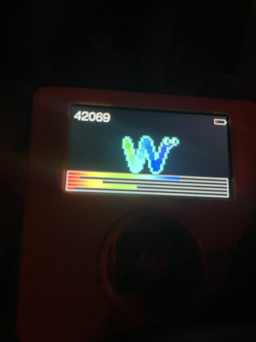 A handheld electronic device displays a colorful waveform-like graphic and the number “42069,” with a battery icon at the top right.