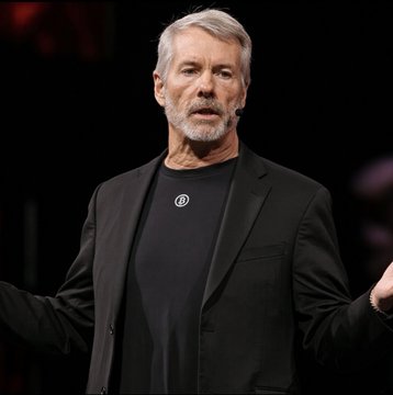 Michael Saylor, wearing a black blazer with a circular pin, speaks on stage while gesturing with his hands, illustrating the post about Strategy ($MSTR) purchasing an additional 334 Bitcoin ($28.7 million) last week as part of its ongoing accumulation strategy.