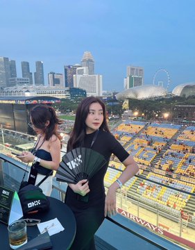 Two young women pose at an outdoor rooftop venue overlooking the Singapore skyline at dusk, with the Marina Bay Sands and Singapore Flyer visible in the background.