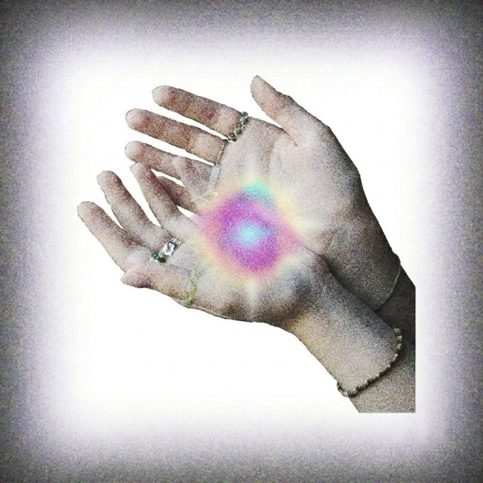 A dreamlike photograph of two hands gently cupped together, featuring silver rings and bracelets, with a glowing iridescent orb of pink, blue, and yellow light emanating from the palms.