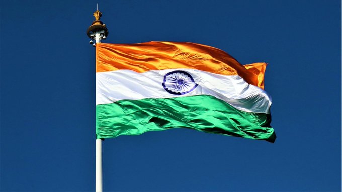 The Indian national flag (saffron, white, and green with the Ashoka Chakra) waves against a clear blue sky from a flagpole.