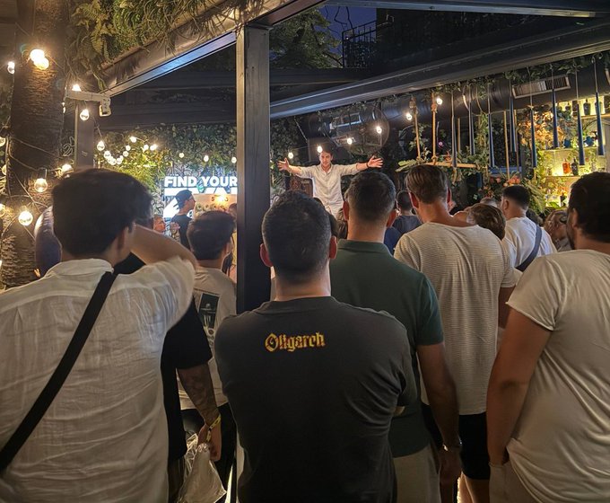 A crowd of people at an outdoor venue watching a speaker on stage under string lights, with a banner reading 'FIND YOUR VOICE' visible in the background.