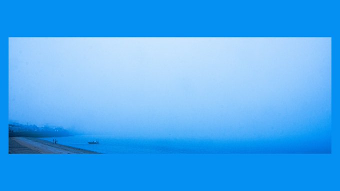 A serene, hazy blue-toned landscape photograph of a coastline or beach, with faint silhouettes of small boats on the water and a distant shoreline under an overcast sky.