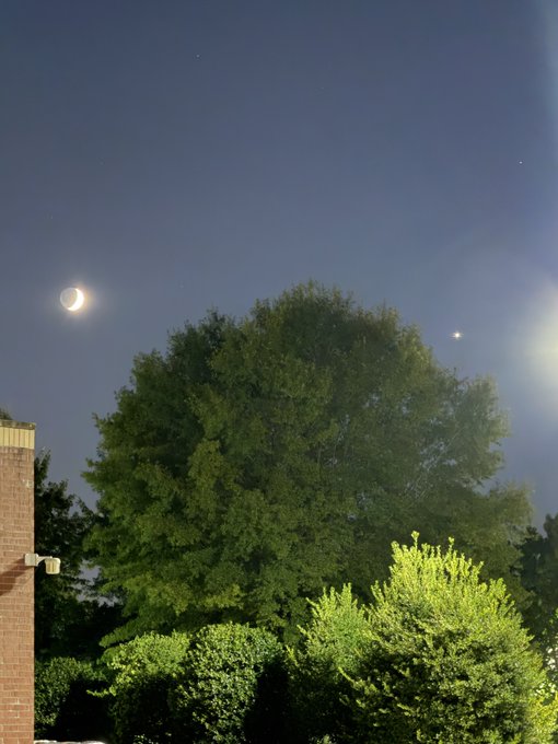 A nighttime photograph showing a crescent moon and a bright star (likely Venus) in a dark blue sky above a large green tree and brick building.