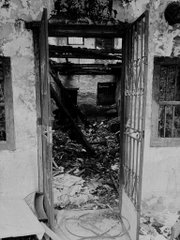 A black and white photograph showing an open doorway revealing a dilapidated, debris-filled interior of an abandoned building.