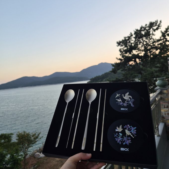 A hand holds a black gift box containing an elegant set of silver-toned spoons and chopsticks with floral coaster designs, set against a coastal landscape at sunset.