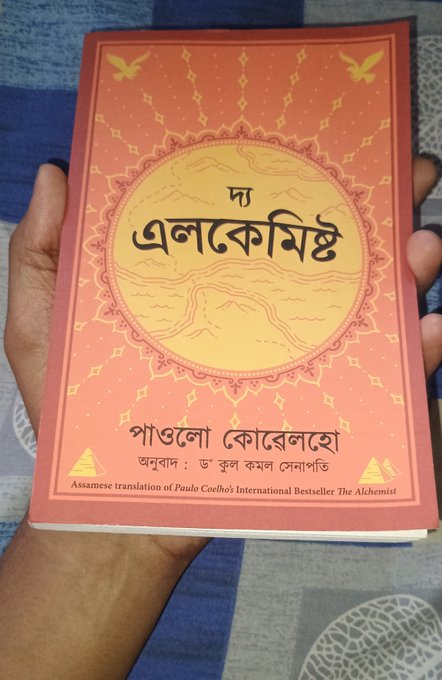 A hand holding a salmon-colored book titled 'Da Alake Mist' in Assamese, which is identified as the Assamese translation of Paulo Coelho's 'The Alchemist'.