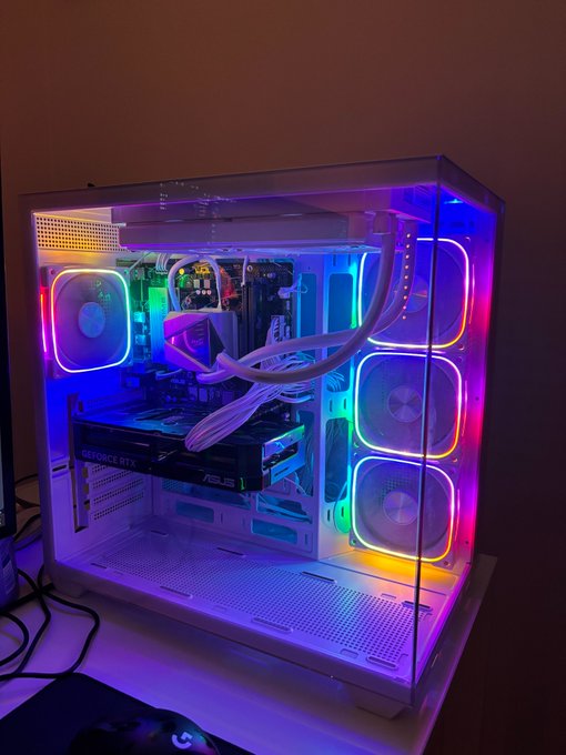 A white PC case with a transparent side panel displaying vibrant RGB rainbow lighting, featuring an ASUS GeForce RTX graphics card and white braided cables.