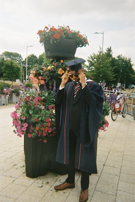A graduate in a black gown and cap adjusts his sunglasses beside a tiered display of colorful flower planters on a paved walkway, with bicycles visible in the background—capturing the traditional milestone moment the caption references before his pivot into crypto.