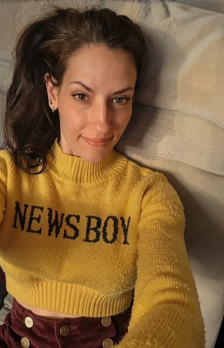 A selfie of a woman with dark hair wearing a yellow sweater with 'NEWSBOY' written across the chest, paired with maroon pants with gold buttons.
