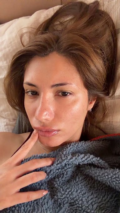 A close-up selfie of a young woman with brown hair lying down, wearing a gray textured top and touching her lip.