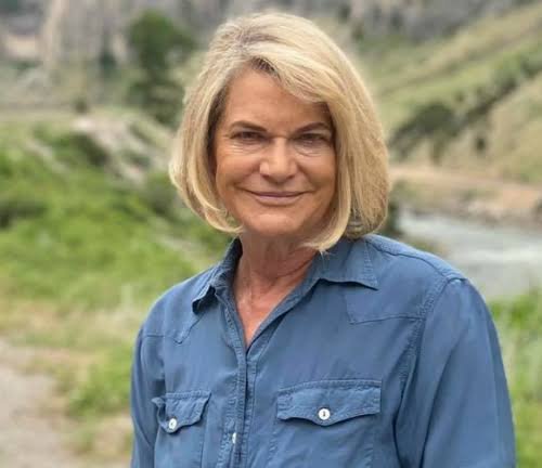 A portrait of an older woman with blonde hair wearing a blue denim shirt, standing outdoors with a natural landscape in the background.
