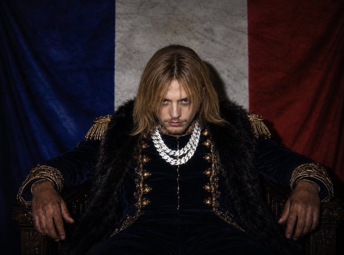 A man with long blonde hair sits on an ornate throne in front of a French flag backdrop, wearing a fur-trimmed jacket adorned with gold details and a large chain necklace.

The image relates to the "Gm @grok and replies guy" post by portraying a regal, Napoleon-like figure, evoking a sense of imperial authority that mirrors the meme's playful reference to engaging with Grok and reply interactions online.