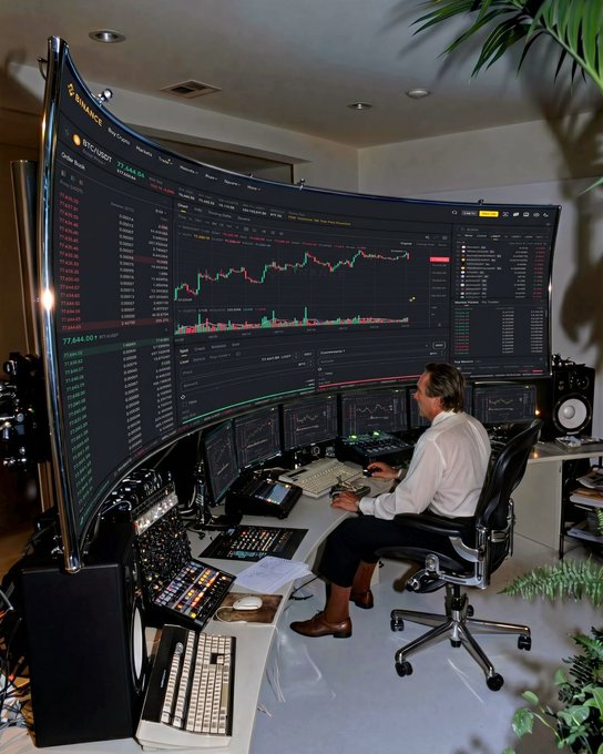 A man sits at an elaborate multi-monitor trading workstation displaying cryptocurrency charts and order books, with the Bitcoin price visible around $77,644.