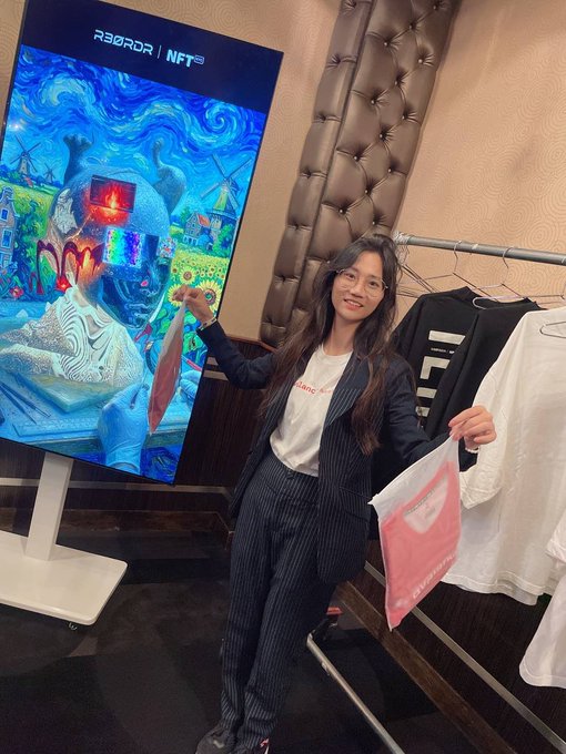 A woman in a pinstripe suit stands next to a digital screen displaying an NFT artwork and 'R3COR | NFT' branding, with clothing items hanging on a rack beside her.