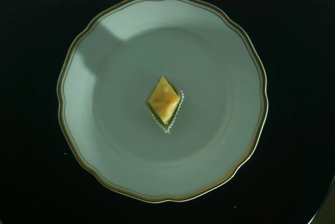A single piece of yellow sushi or food item sits in the center of an empty white plate against a dark background.