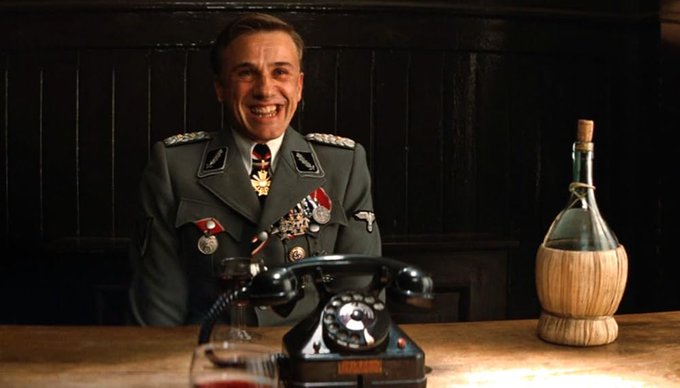 The image shows a man dressed in military uniform sitting behind a desk with an old-fashioned phone, smiling at the camera as if posing for a photo. In the background, there's an old bottle of wine, which appears to be uncorked. The price of Bitcoin is mentioned in the post above the picture. However, it doesn't seem directly related to the man or his position behind the desk, but rather, it seems to serve as background information about cryptocurrency.