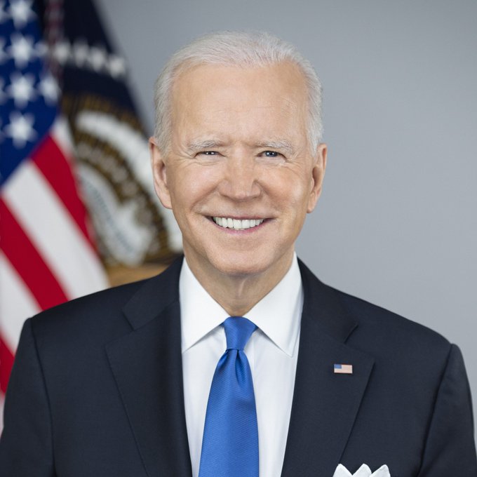 A man in a dark suit with a blue tie, smiling broadly with an American flag in the background, appears to resemble former U.S. President Joe Biden. The image relates to the post about his son Hunter Biden launching a memecoin called $LAPTOP, likely serving as a recognizable reference to the Biden family amid the news.