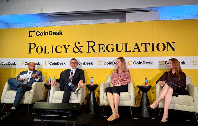 Four panelists sit in armchairs on a stage at a 'CoinDesk POLICY & REGULATION' event, with a bright yellow branded backdrop behind them.