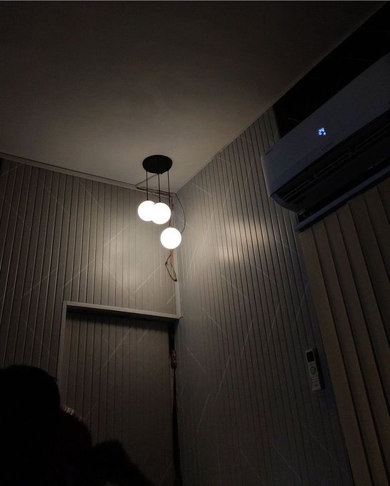 A dimly lit room interior featuring a modern pendant light with three white globe bulbs hanging from a black ceiling fixture, mounted on walls with vertical paneled wallpaper, with an air conditioner visible in the corner.