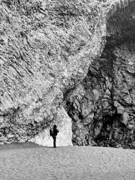 A black-and-white photograph showing a tiny person standing at the entrance of a massive rocky gorge or cave, emphasizing the dramatic scale of the stone cliffs.