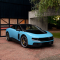 A light blue Lamborghini sports car parked on a brick driveway with greenery and a building in the background.