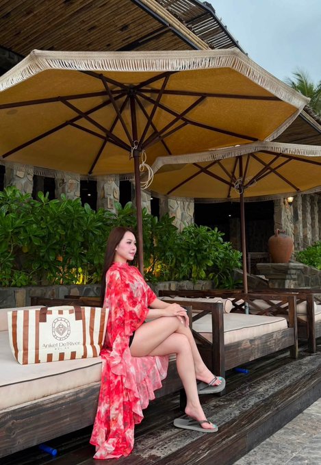 A woman in a flowing red floral dress sits on a wooden bench under a tan umbrella at the Ayodya Resort, with a matching striped tote bag beside her.