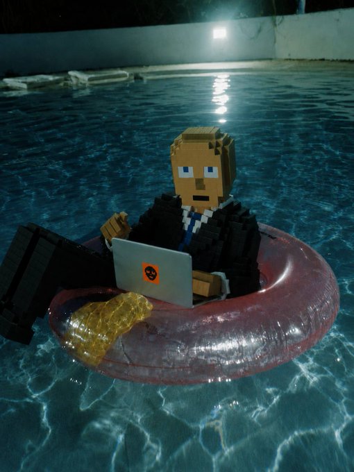A blocky, pixelated character in a black suit lounges on a pink inflatable floatie in a nighttime pool, working on a laptop marked with an orange skull logo. The image humorously depicts the post's theme of "working on some things" while floating leisurely, with the skull logo reinforcing the mysterious, edgy tone of the cryptic update.