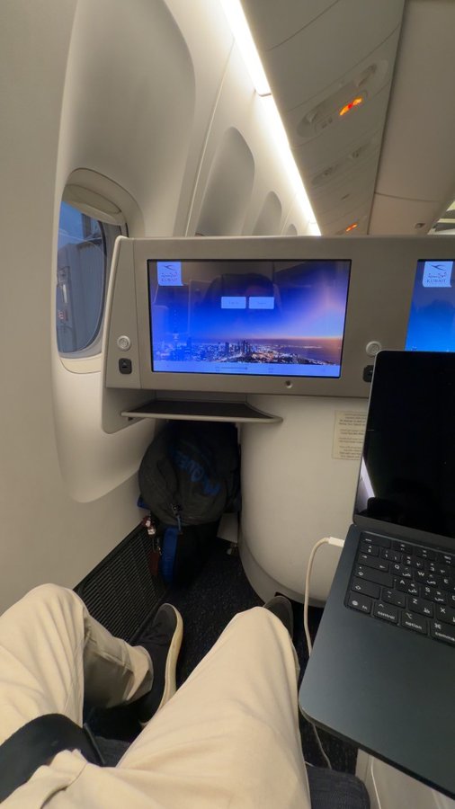 A first-person view from an airplane window seat showing the passenger's legs, a backpack on the floor, and an in-flight entertainment screen displaying a view above the clouds with 'Return' and 'Next' buttons.